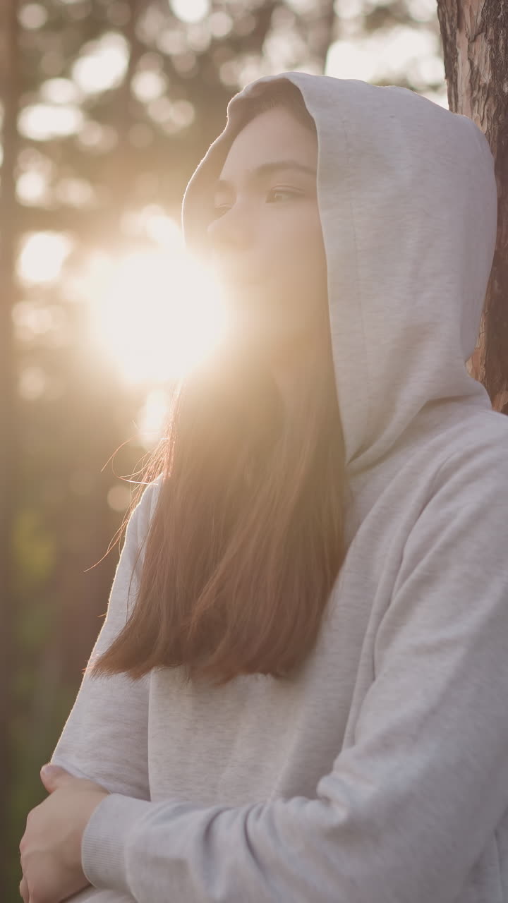 дама в капюшоне отдыхает, опираясь на дерево в парке. женщина думает одна о депрессии и одиночестве, гуляя на свежем воздухе вечером. подавленные эмоции