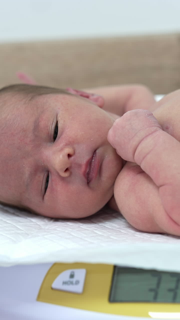 Naked child lying on the electronic scales and hiccupping. Cute Newborn baby being weighed in Maternity unit in the hospital. Vertical video