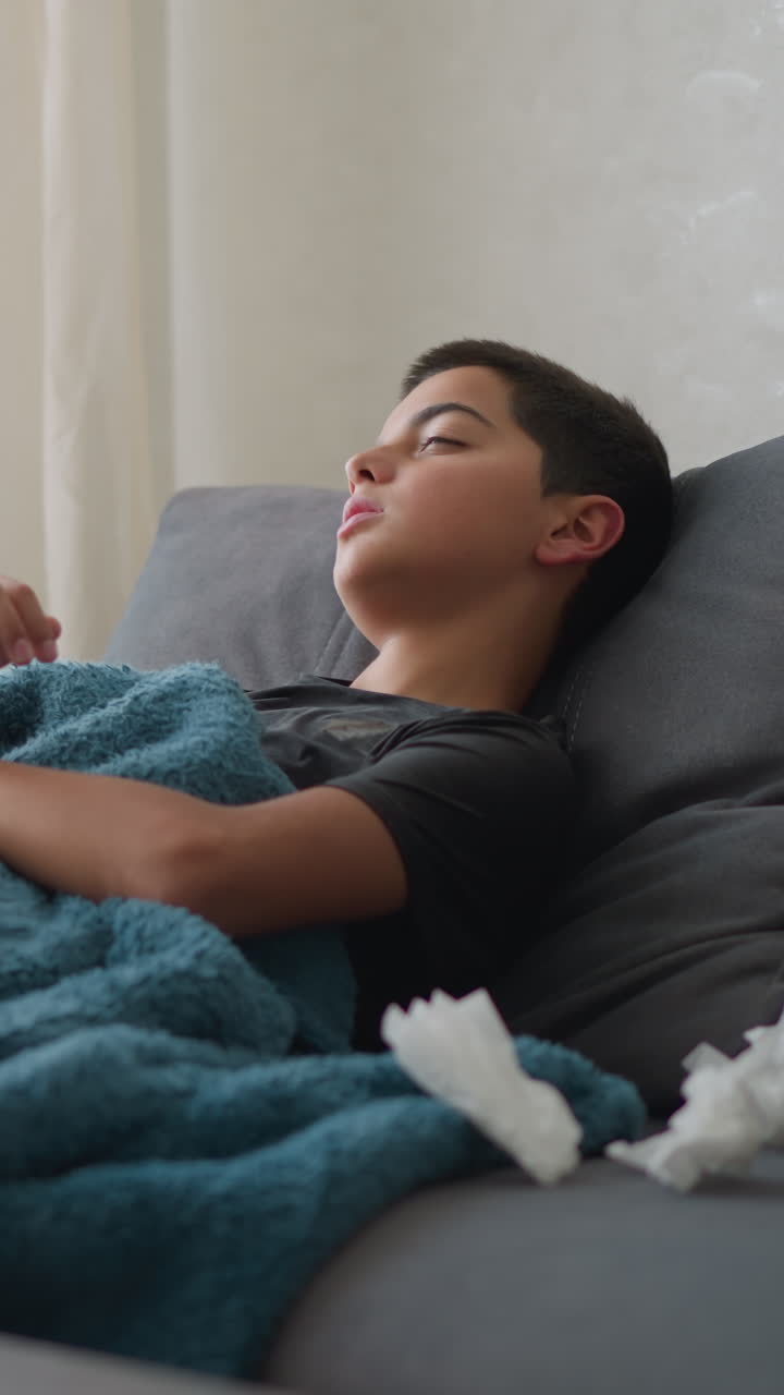 Boy sleeping on couch covered in blanket adjusting for comfort, tissues nearby and glass cup on table, relaxing indoors with natural light streaming through window