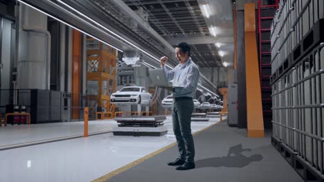 vista lateral completa del cuerpo de un trabajador profesional masculino asiático de pie con su portátil en la fábrica de automóviles ev, instalación industrial de alta tecnología, levanta el puño con un objetivo gritón después de escribir en la portátil