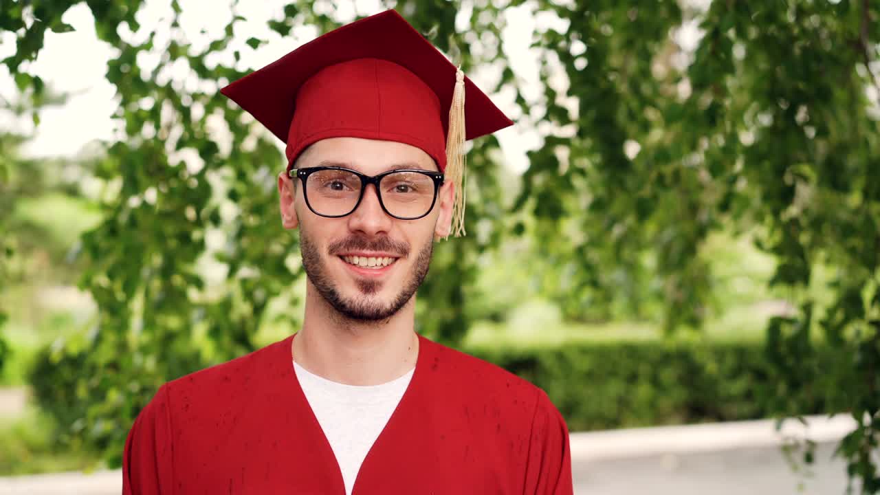 Portrait of bearded young man graduating student wearing glasses, graduation gown and mortar-board smiling and looking at camera standing outdoors. People and education concept.