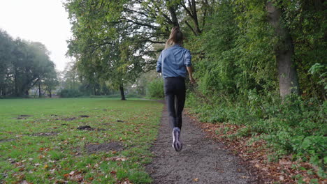 dynamic slow motion shot: camera follows active woman running through park on path, forest on right, grassfield on left