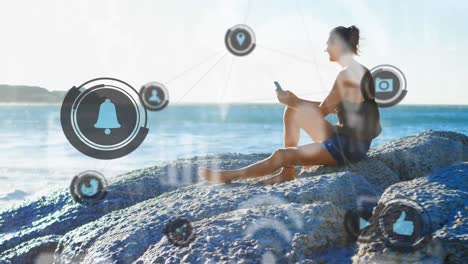 red de iconos digitales contra una mujer caucásica que usa un teléfono inteligente sentada en las rocas cerca del mar