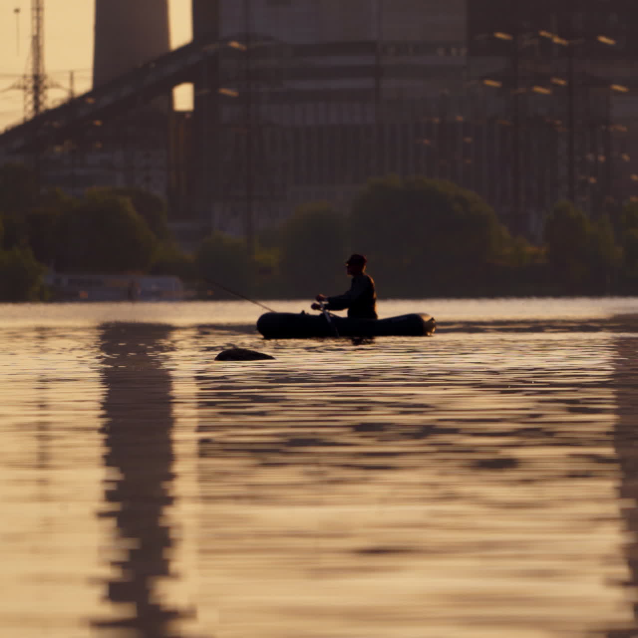 Fisherman in a boat with fishing rods in the evening. Man in hat floating in a boat and fishing on the background of a factory in the city.