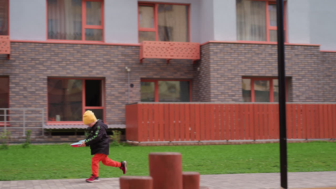 Child in yellow hat and dark jacket runs through playground holding toy boat, surrounded by benches, modern apartment buildings, and soft natural light, showing joyful outdoor momen