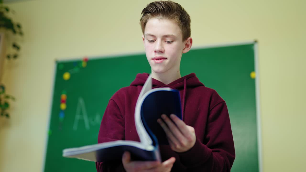 Boy looking into the school journal. Teenager standing in the classroom and reading information in the folder. Learning process at school.