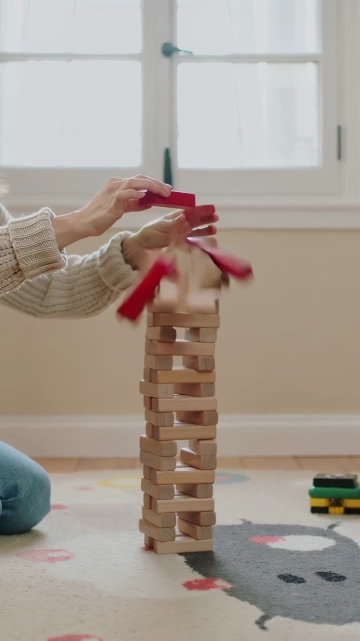 Video verticale: Aggiungendo un blocco rosso, un adulto e un bambino con una maglietta blu a maglia impilano una torre su un tappeto da gioco, facendola poi cadere.