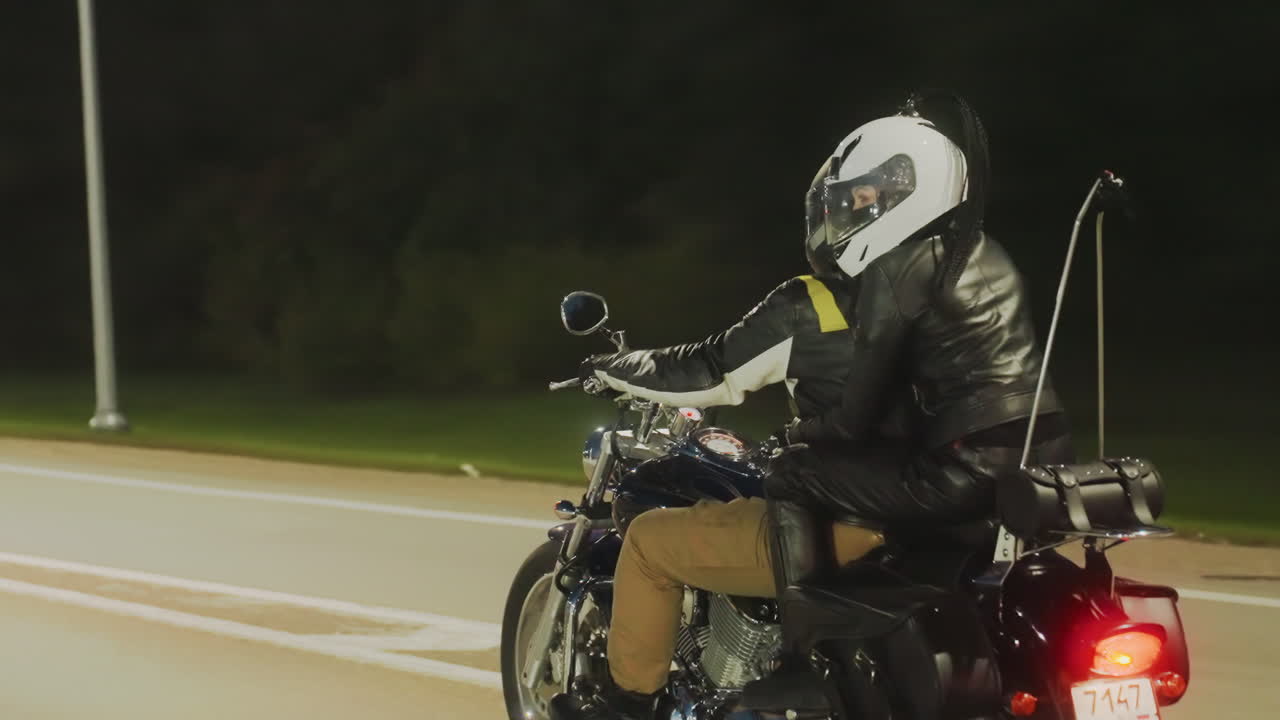 Riders on motorcycle road trip pass pedestrian signpost and roadside shelter during night journey, illuminated by street lights, wearing helmets and leather jackets
