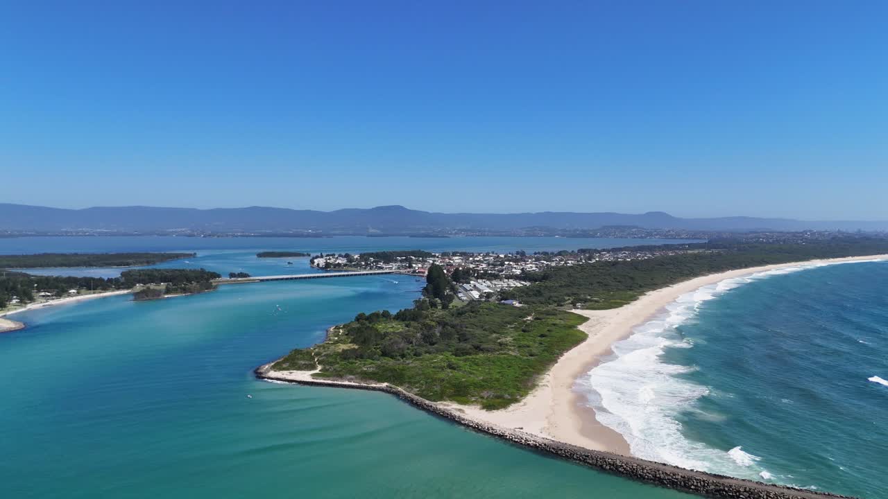 Vista panoramica aerea di Windang Inlet con lingua di sabbia, canale blu intenso e onde che incontrano acque calme, cielo blu vivace