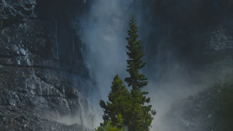 a cinematic slow motion shot of huge pine trees and a the huge and stunning takakkaw waterfall in the background