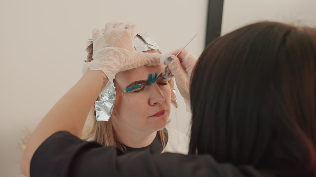 Nahaufnahme einer Frau, die sich die Augenbrauen über die Schulter färbt, Stylist formt mit behandschuhten Händen und Folienakzenten türkisfarbenen Farbstoff über die Brauen, Kundin reagiert mit konzentriertem Gesichtsausdruck, modernes Saloninterieur, aufmerksam