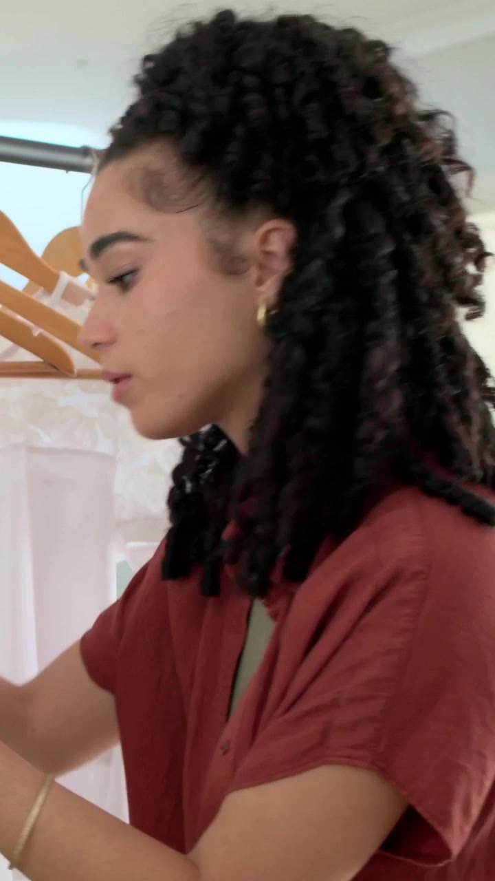 Vertical video: Approaching rack, woman in rust shirt inspecting garments at home, selecting outfit