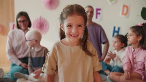 una niña feliz de preescolar con cabello rubio en una camiseta amarilla mira a la cámara y sonríe en el fondo de su primera lección con los maestros en las clases de escuela introductoria. retrato de una niña pequeña mirando a la cámara en el telón de fondo de su primer lección con los maestros