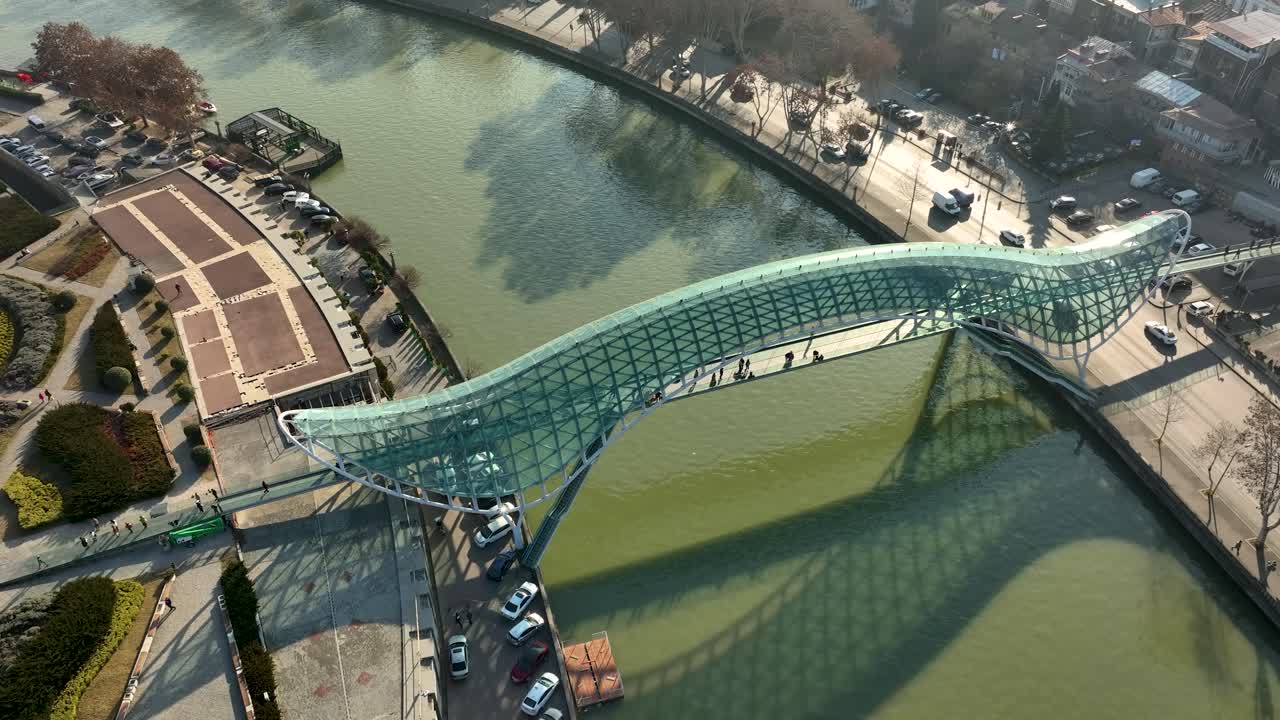 Drone shot of the Bridge of Peace in Tbilisi, Georgia, crossing the green Kura River. The bridge’s futuristic structure stands out with sunlight and urban surroundings