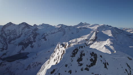 Drone-flying-by-top-of-snowy-Kitzsteinhorn-mountain-with-big-cross-on-it-revealing-mountain-range-on-sunny-day