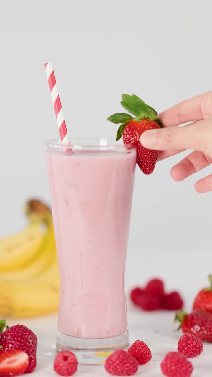 Una mano guarnisce un frullato rosa cremoso con una fragola fresca, circondato da frutta sotto una luce brillante.