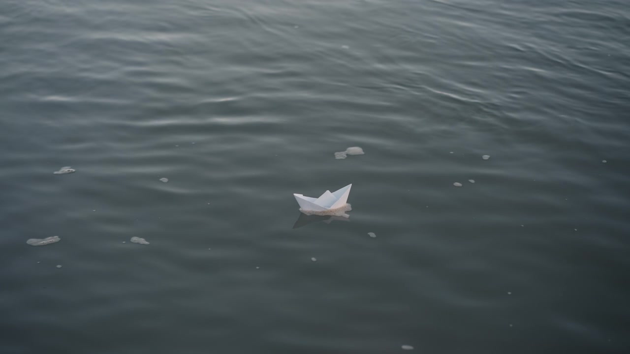 White paper boat gently drifts on calm water surrounded by small floating debris. scene features soft natural light and tranquil aquatic environment with subtle ripples and reflections