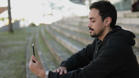 Side-view-of-young-Arabic-man-with-dark-curly-hair-and-beard-in-black-hoodie-sitting-on-stairs-outside