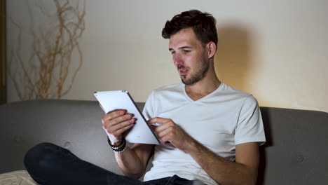 A-young-man-with-a-stubble-and-a-modern-hairstyle,-wearing-a-white-t-shirt-and-jeans,-sitting-at-home-on-a-sofa,-legs-crossed,-swiping-on-his-tablet-while-browsing-the-internet,-static-4k