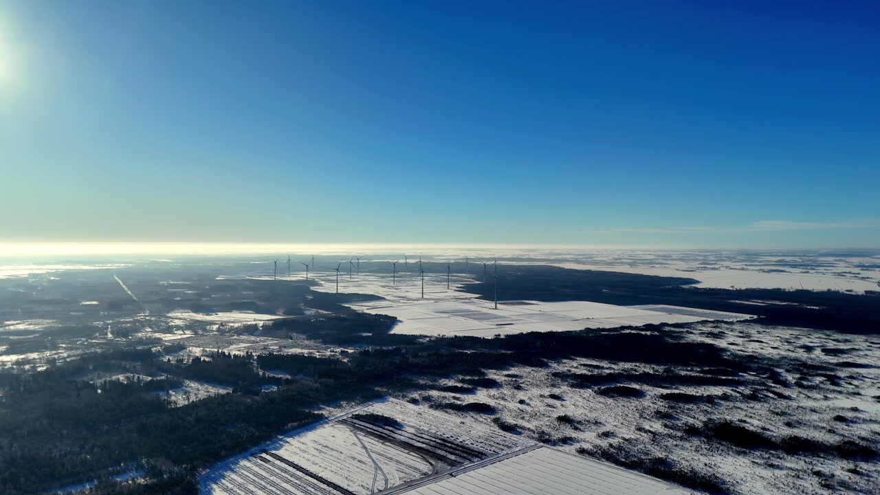 澄み切った青空の下、柔らかな朝の光を浴び、遠くの地平線に霧がかかる広大な雪に覆われた冬の風景にそびえ立つ雄大な風力タービンを、高高度から空撮