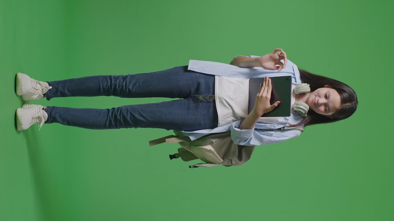 cuerpo lleno de estudiante adolescente asiática con una mochila sonriendo y usando una tableta mientras está de pie en el estudio de fondo de la pantalla verde
