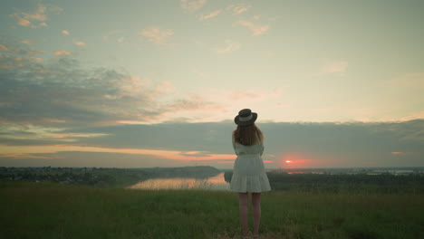 back view of a wife in a white dress and hat, arms folded, standing alone in a serene grassy field, gazing at a tranquil lake during sunset. the scene is calm and peaceful colors in the sky