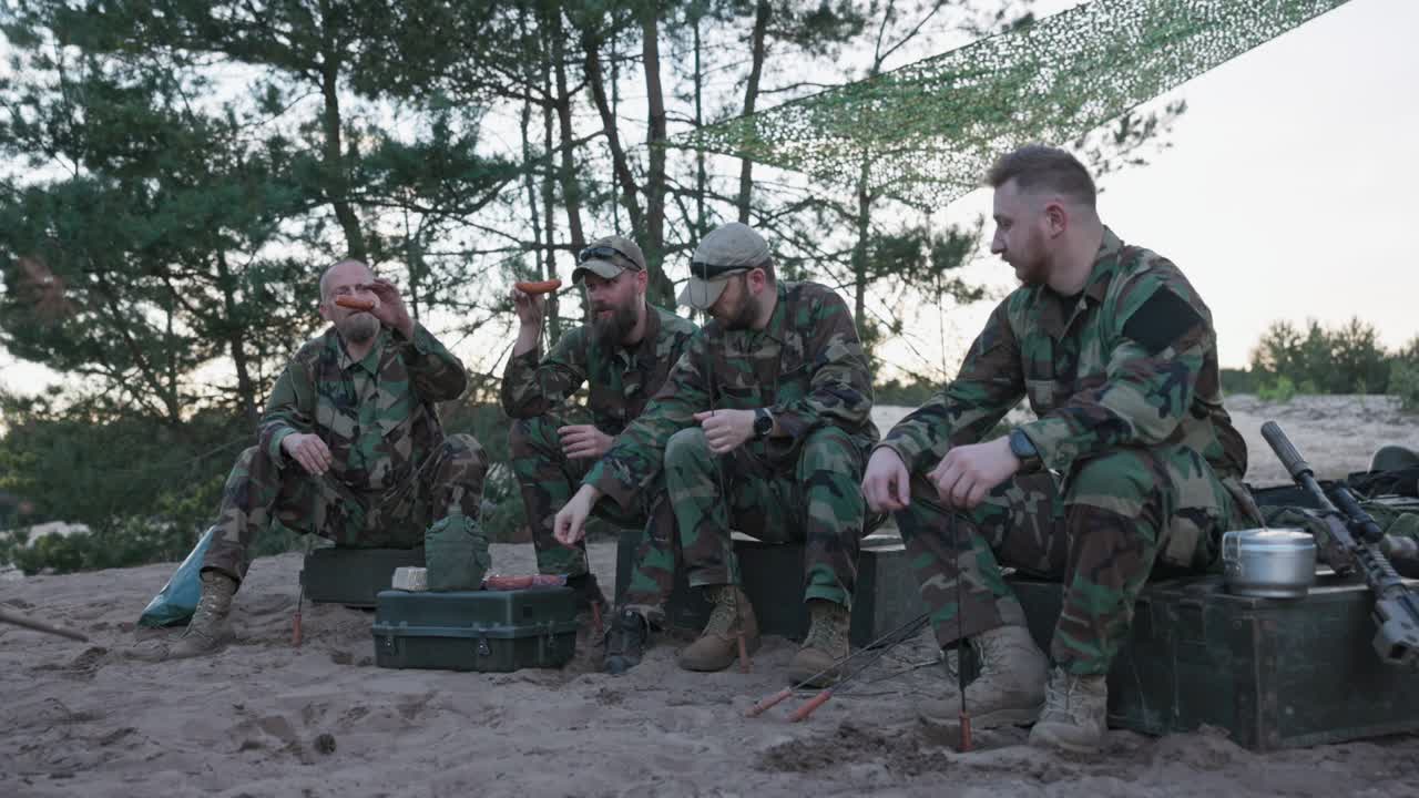 hombres militares en uniformes moro relajándose después de un turno de la tarde en la base en el campo, sentados en cajas afuera y preparando una fogata, recolectando salchichas en palos, preparando la cena