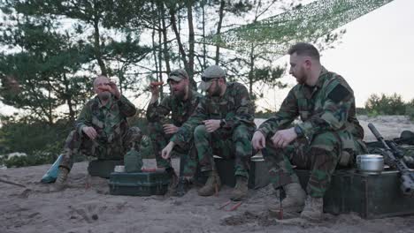 military men in moro uniforms relaxing after an afternoon shift at the base in the field, sitting on crates outside and preparing a campfire, scooping sausage on sticks, preparing dinner