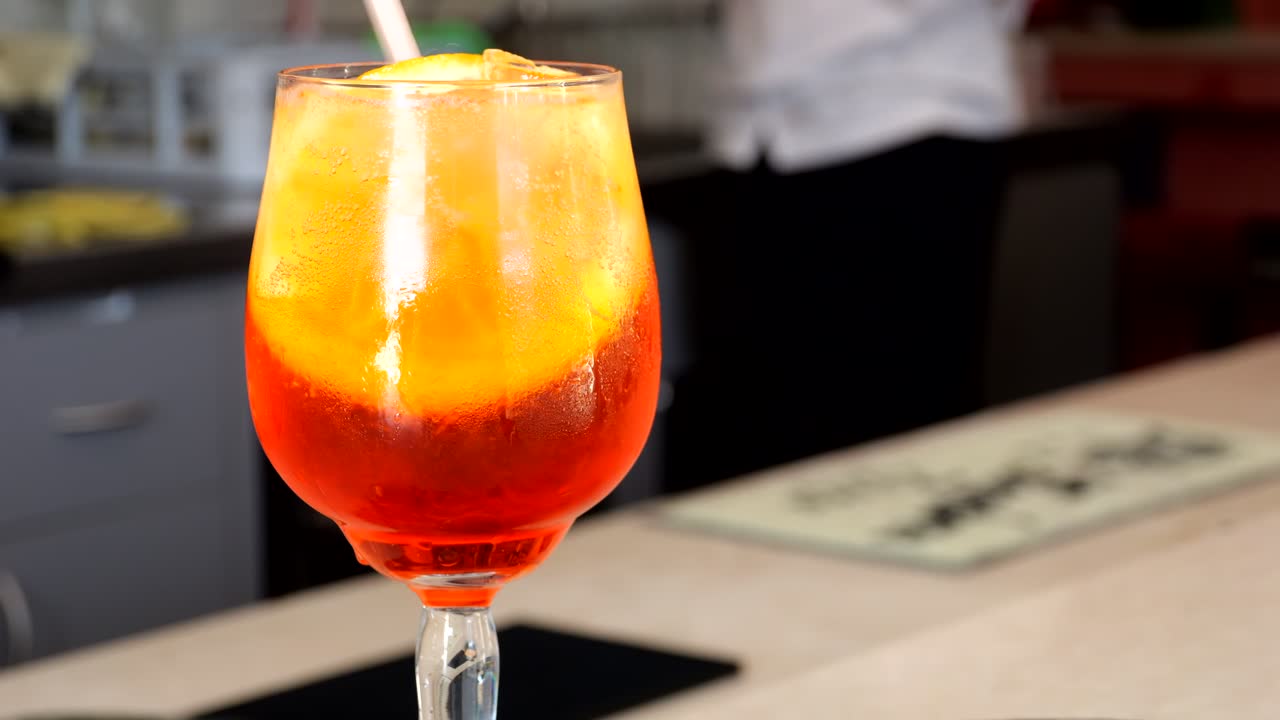 Cocktailglas fyldt med en levende orange cocktail, isterninger og en citrusskive står på en bardisk i afslappede indendørs omgivelser, fanget i et statisk nærbillede