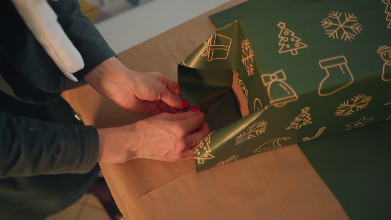 Closeup woman folding gift wrap corner on green patterned paper with precise hand movements and red nail polish, tape and scissors nearby, calm focused craft moment, finishing touches for holiday