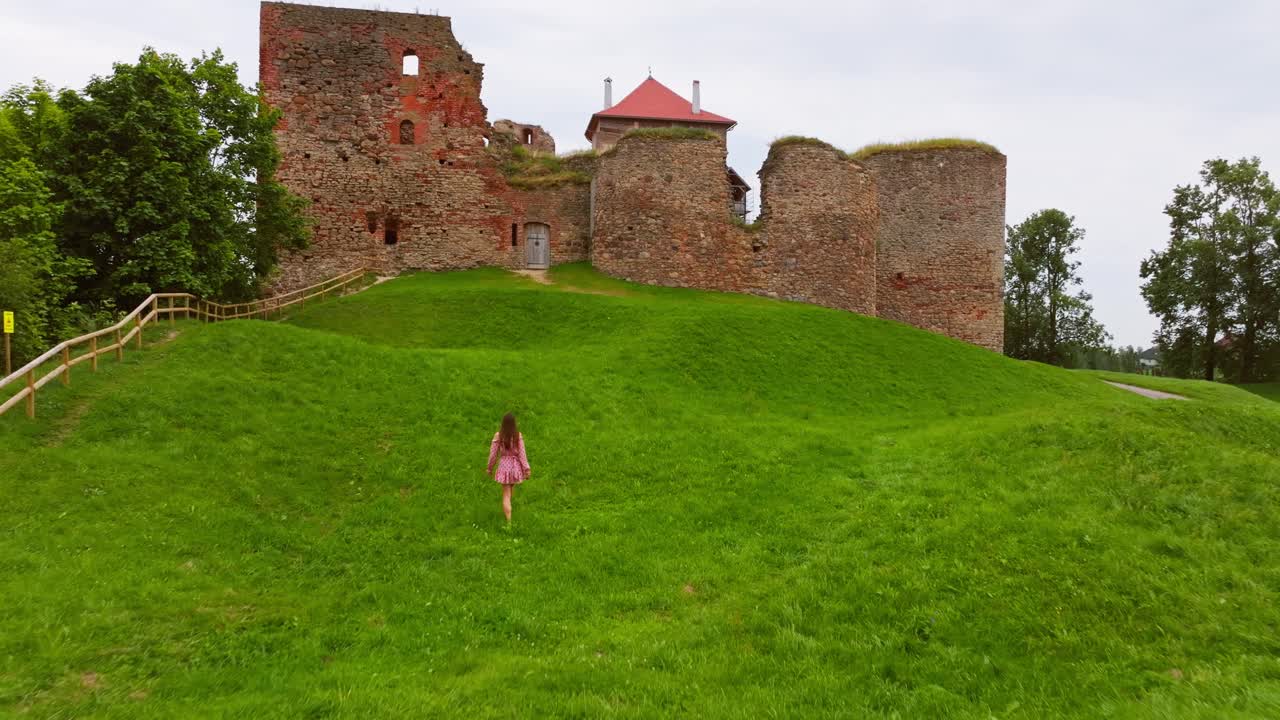 Woman in pink dress climbs hill toward Bauska Castle ruins, overcast summer day