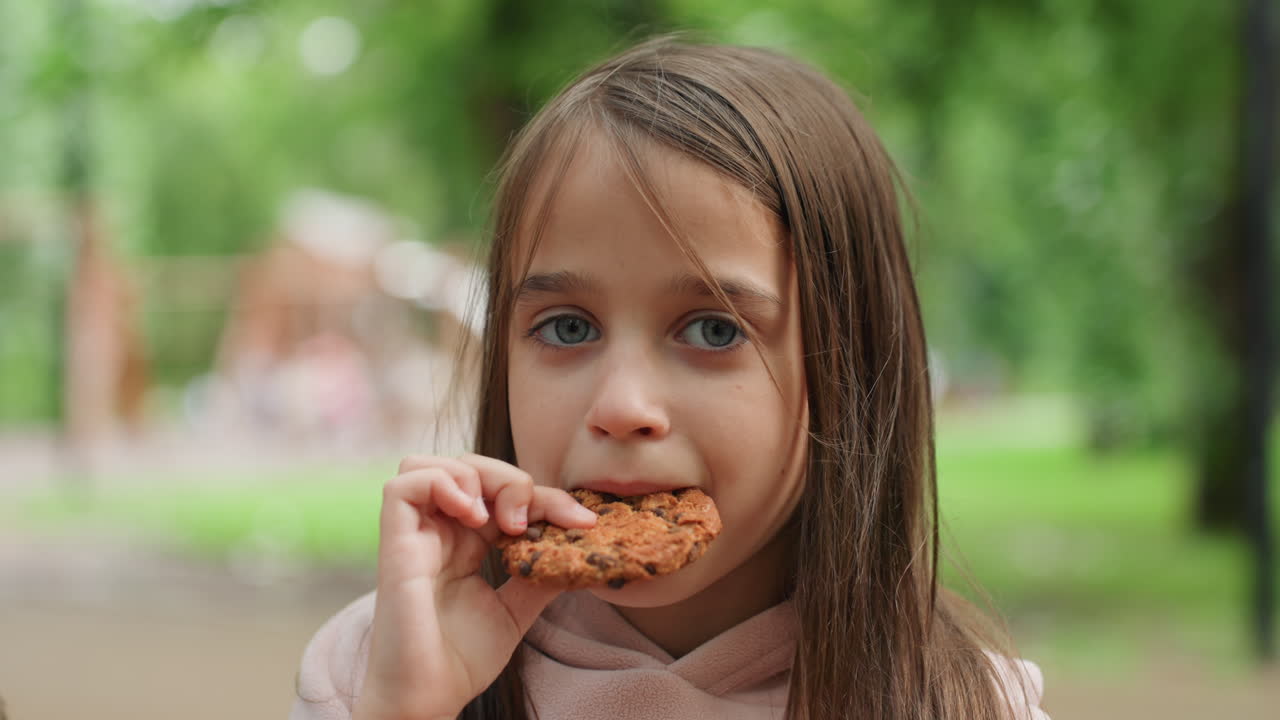 Young daughter eating cookie in park, closeup portrait capturing quiet candid moment with braided hair, soft bokeh background of trees and playground, warm afternoon snack, innocent gaze and gentle