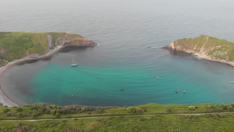 an aerial sideway (left to right) footage of the lulworth cove with sandy beach, crystal blue water, fishing boat, kayakistes and crashing waves on the rocky cliff