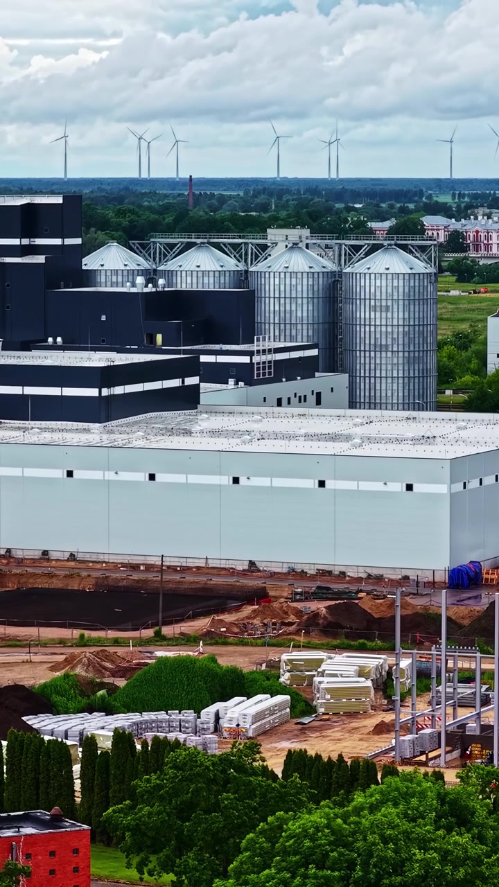 Store industribygninger og metalsiloer står ved siden af et aktivt byggepladsområde, med vindmøller synlige i det fjerne landskab - Luftoptagelse der daler ned