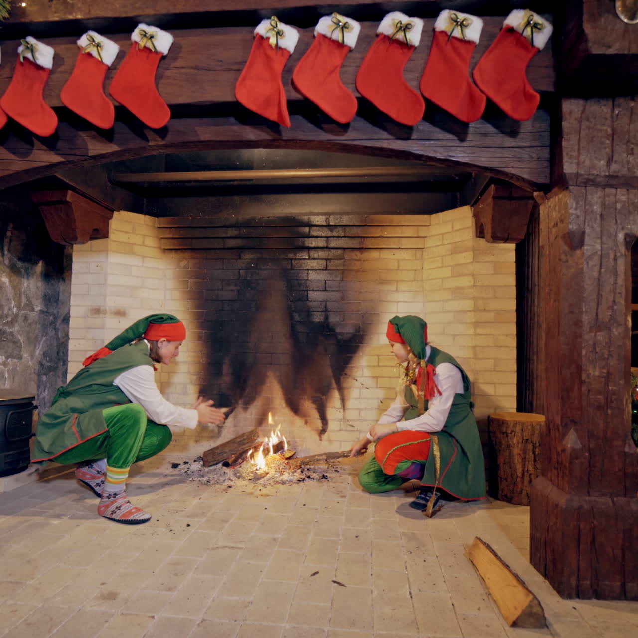 Elves near fireplace on Christmas background indoors. Funny elves in green costumes sit near the fire in the house.