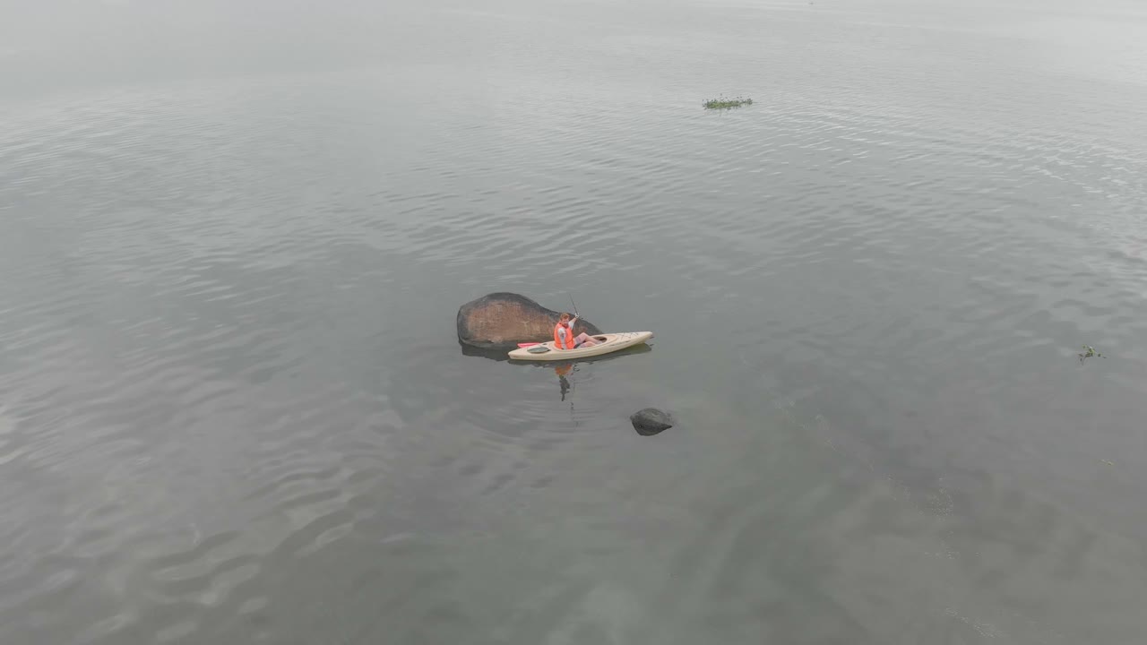 Aerial orbiting around a man fishing from a kayak on Lake Victoria near a granite bolder
