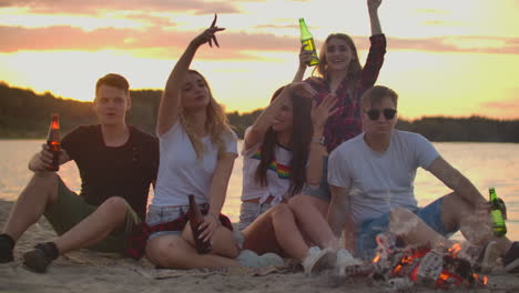 the young colleagues are sitting in around bonfire on the lake coast. they are dancing with beer and talking to each other at sunset.