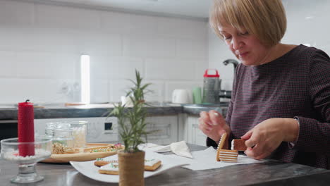 close-up of woman decorating cookies to create house-shaped design with icing on kitchen table filled with flower cookies, baking essentials, and a potted plant in cozy kitchen setting