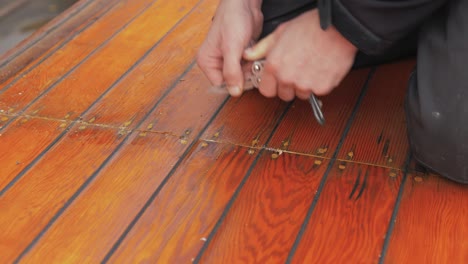 young man uses pocket knife to clean out old mastic of wooden boat roof planking close up