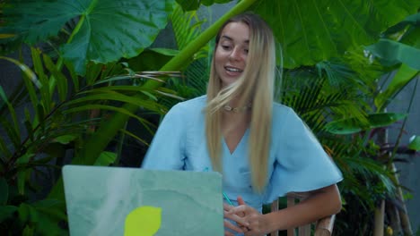 a girl in a blue dress remotely online working behind a laptop and looking into the screen the backyard with green plants