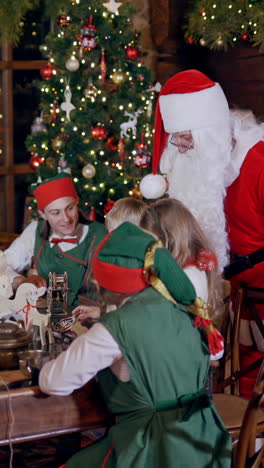 santa claus, elves in green costumes and two kids preparing gifts and letters for holiday. elves and children are sitting at the wooden table on the background of christmas decorations. vertical video