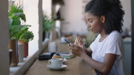 Feliz-Mujer-Afroamericana-Usando-Un-Teléfono-Inteligente-En-Una-Cafetería-Navegando-Mensajes-En-Línea-Disfrutando-De-Compartir-El-Estilo-De-Vida-En-Las-Redes-Sociales-Relajándose-En-Un-Restaurante-Cafetería