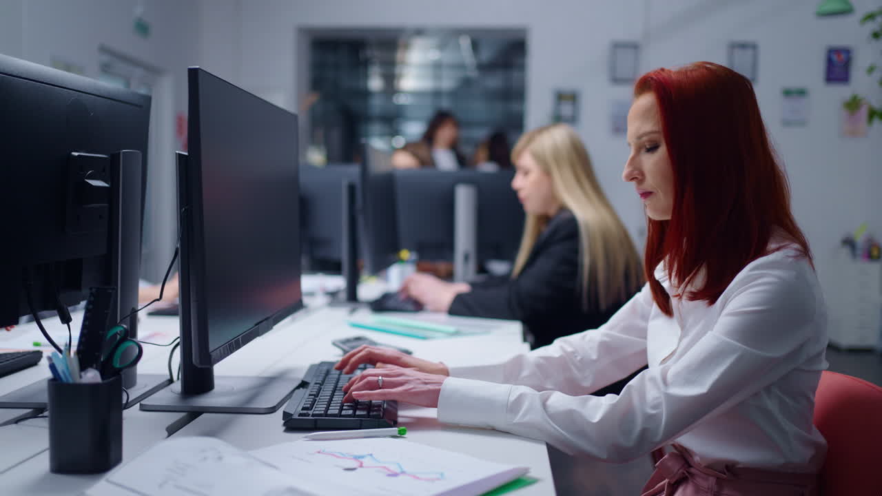 A woman with red hair works on a computer in an office