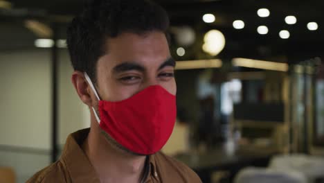 Portrait-of-a-mixed-race-businessman-wearing-face-mask-looking-at-camera-in-modern-office