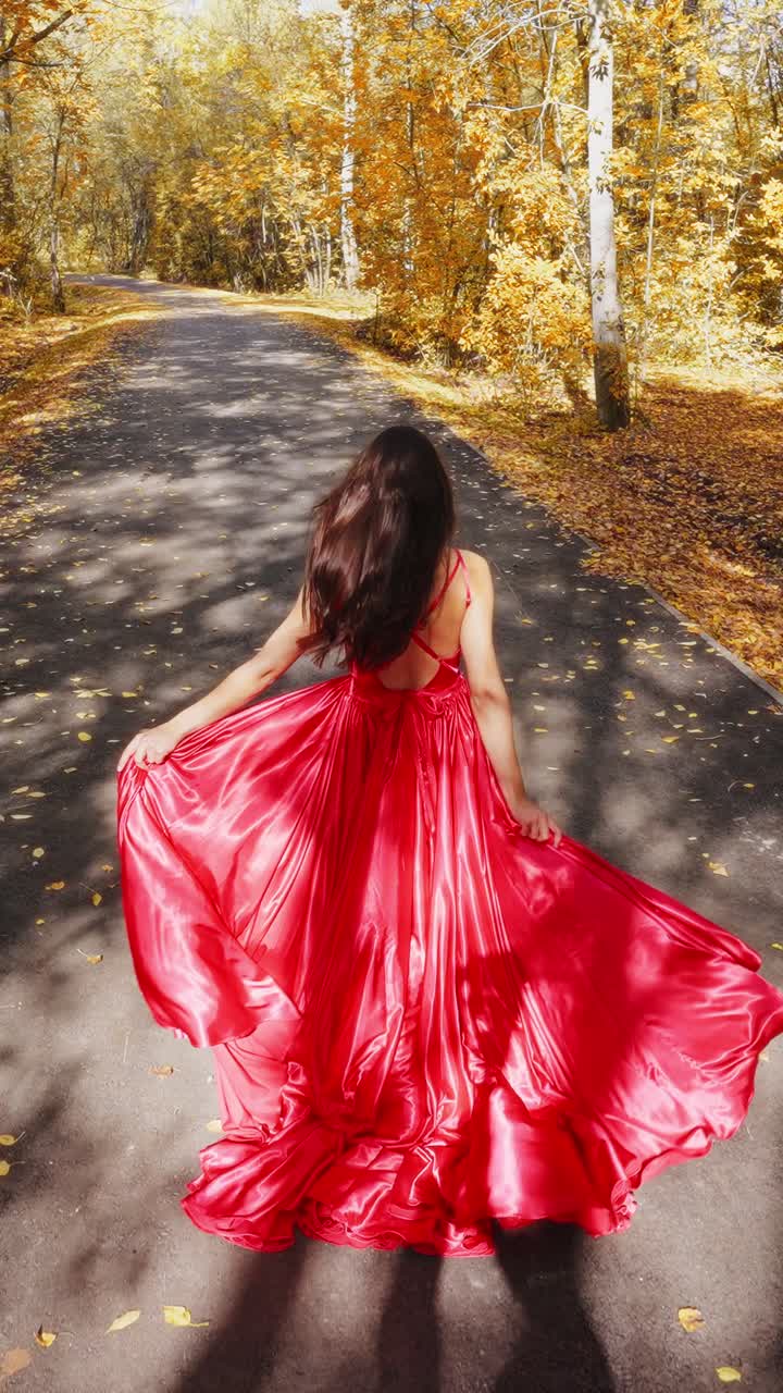 A Woman in a Flowing Red Dress Walks Along a Leaf-Covered Autumn Path, Surrounded by Golden Foliage, Capturing the Essence of Fall's Vibrant Colors and Natural Beauty