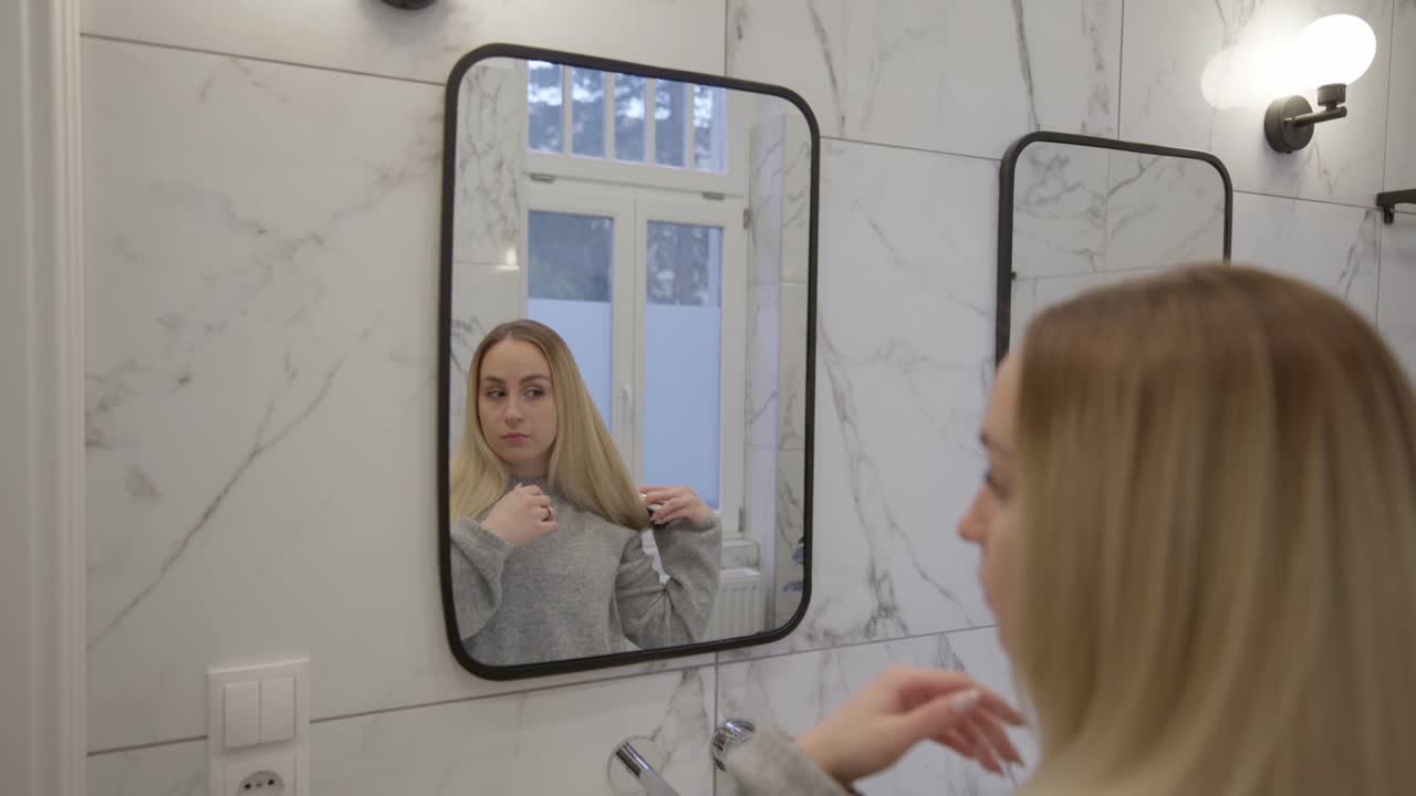 Una giovane donna si trova in un bagno moderno, guardando il suo riflesso nello specchio mentre si sistema i capelli.