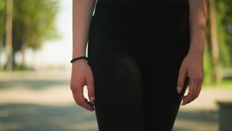 lower body view of lady in black athletic wear walking outdoors with black hand bangle, featuring close-up of arm and blurred background of lush greenery, under warm sunlight