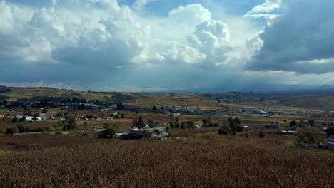 Aerial-view-establishing-of-corn-fields-with-a-cloudy-landscape-with-sun-rays,-several-separate-houses-with-lots-of-space,-mountains-of-Almoloya-Mexico