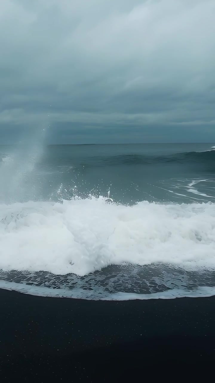 Vertical video: Rising swell pushing breaking surf onto black sand beach, sending white foam spray