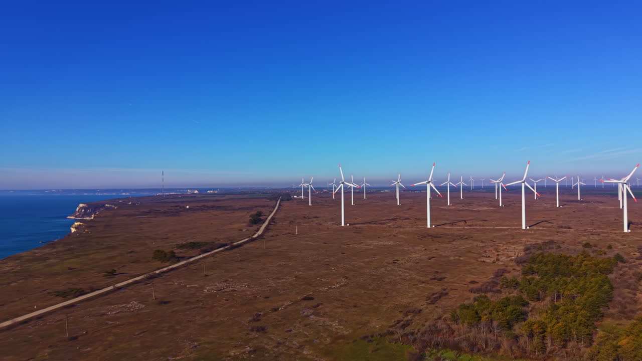 As turbinas eólicas giram em um dia claro perto de uma área costeira com terreno aberto e uma estrada sinuosa ao fundo, mostrando os esforços de energia renovável e o belo terreno